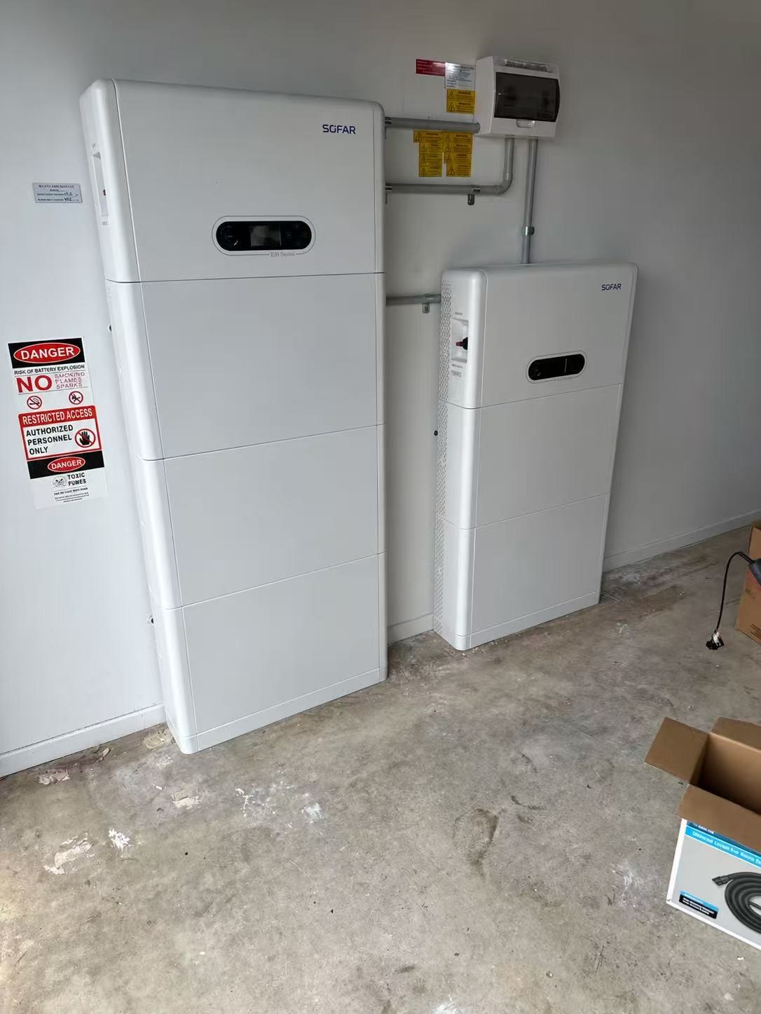 An E3 Energy SOFAR installation: a tall SOFAR stack with an ESI inverter display on top and a shorter SOFAR stack beside it, mounted against a garage wall under the switchboard